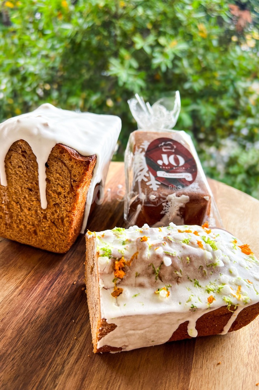 Trois cakes glacés, dont un partiellement tranché, posés sur une planche en bois devant un fond végétal.
