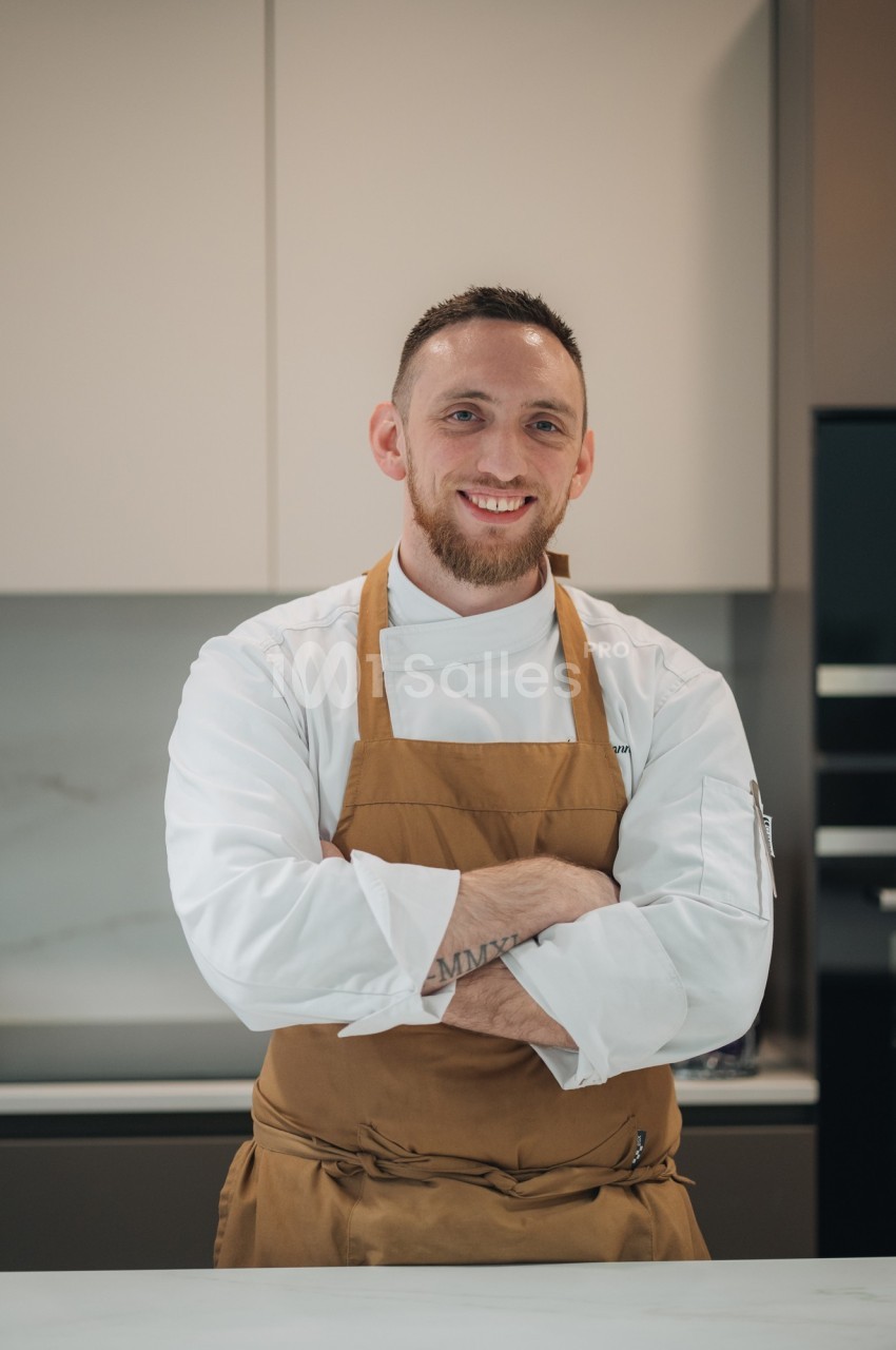 Un chef en tablier marron croise les bras devant un plan de travail dans une cuisine moderne.