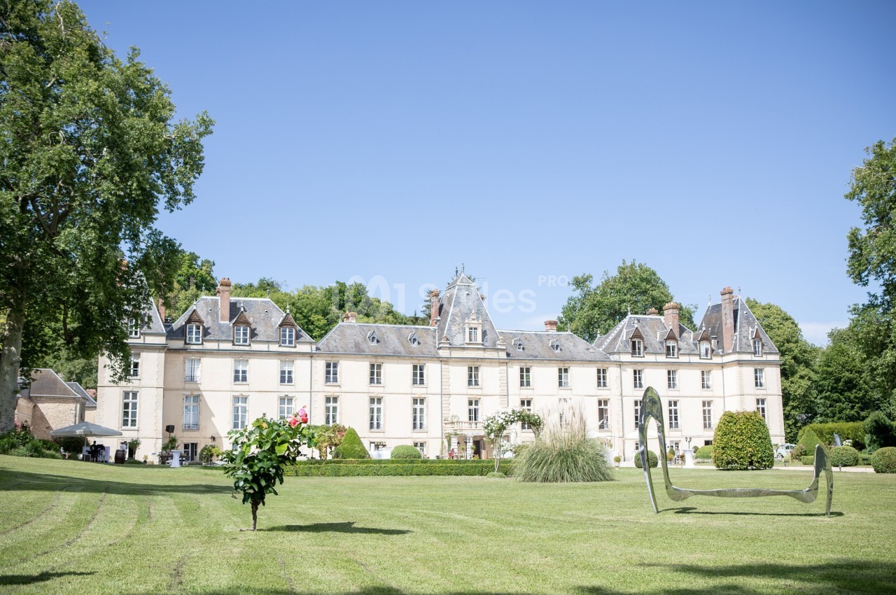 Façade d'un château entouré d'un parc verdoyant avec pelouse, arbres et sculptures modernes.