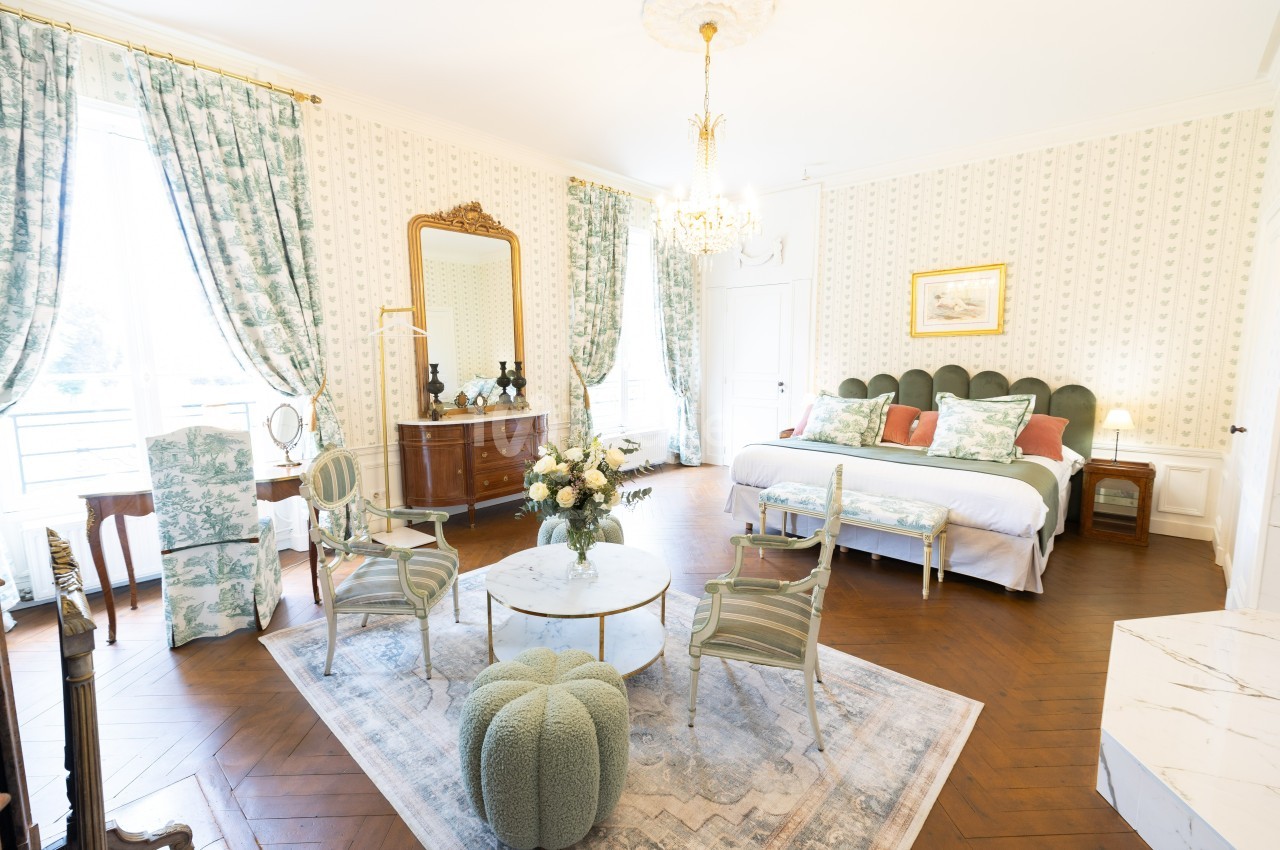 Chambre lumineuse avec lit double, meubles anciens, fauteuils, table basse et rideaux à motifs floraux.