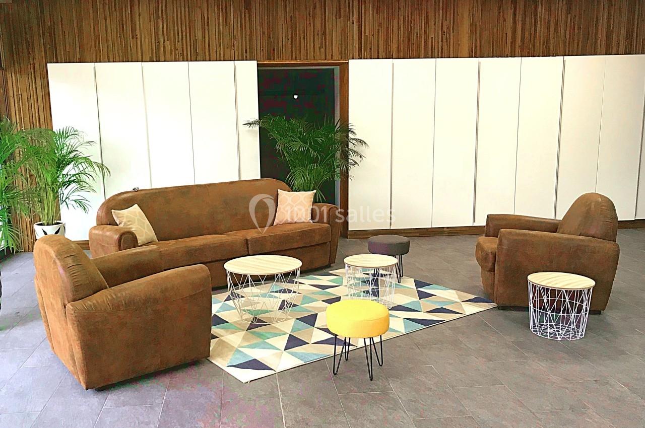 Salon avec canapé, fauteuils en tissu brun, tables basses géométriques et plantes vertes dans un espace lumineux.