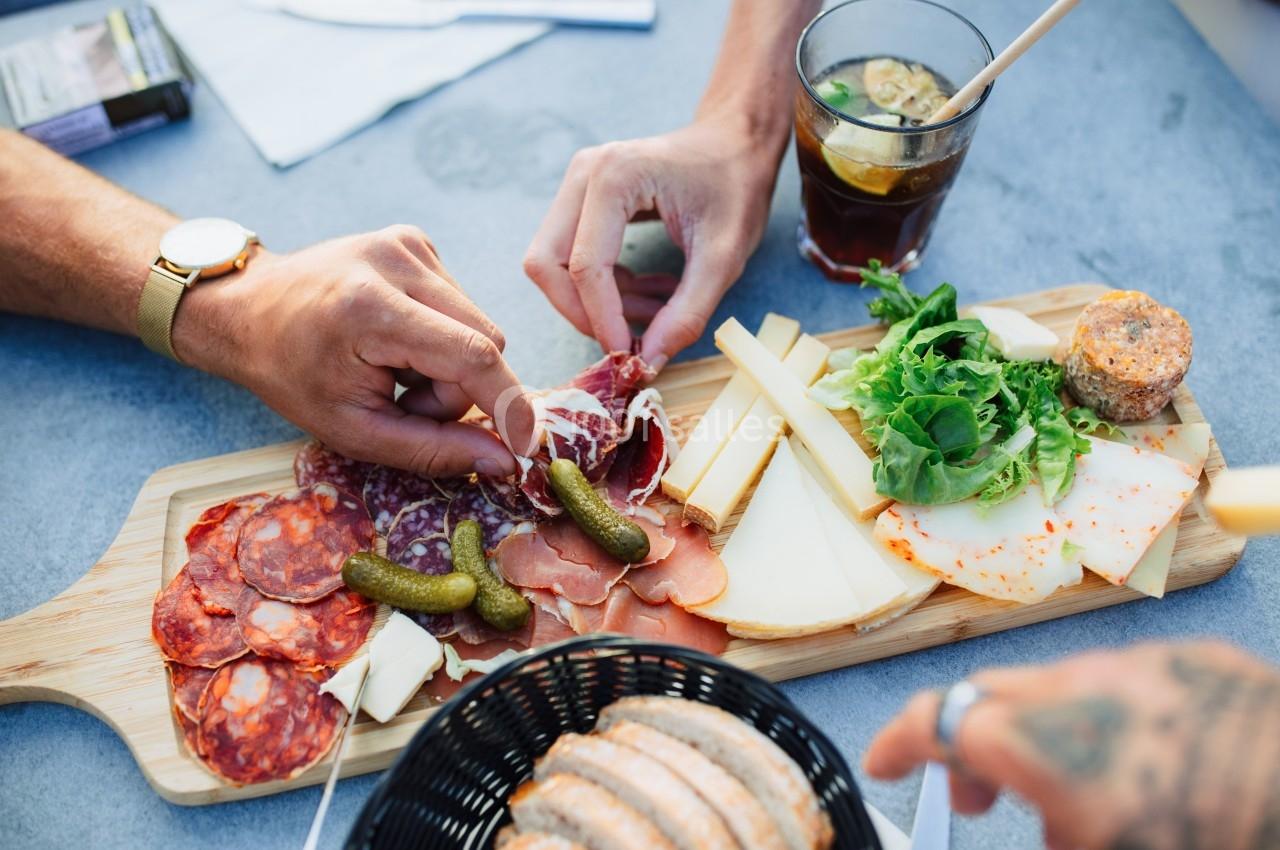 Planche de charcuterie et fromages variés avec cornichons, salade et pain, servie sur une table avec des boissons.