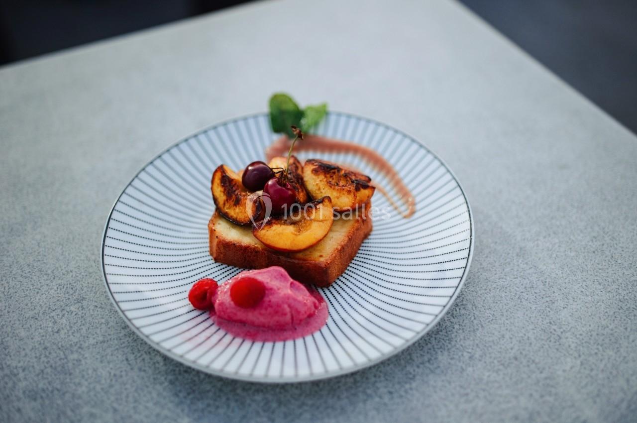 Tranche de gâteau garnie de pêches caramélisées, cerises, framboises et crème rose, servie sur une assiette rayée.