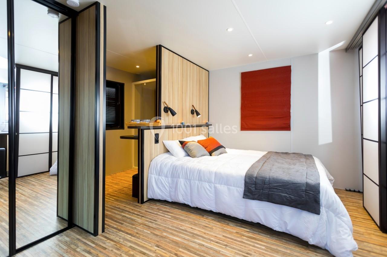 Chambre moderne avec lit double, parquet clair, cloison en bois, stores rouges et éclairage intégré.