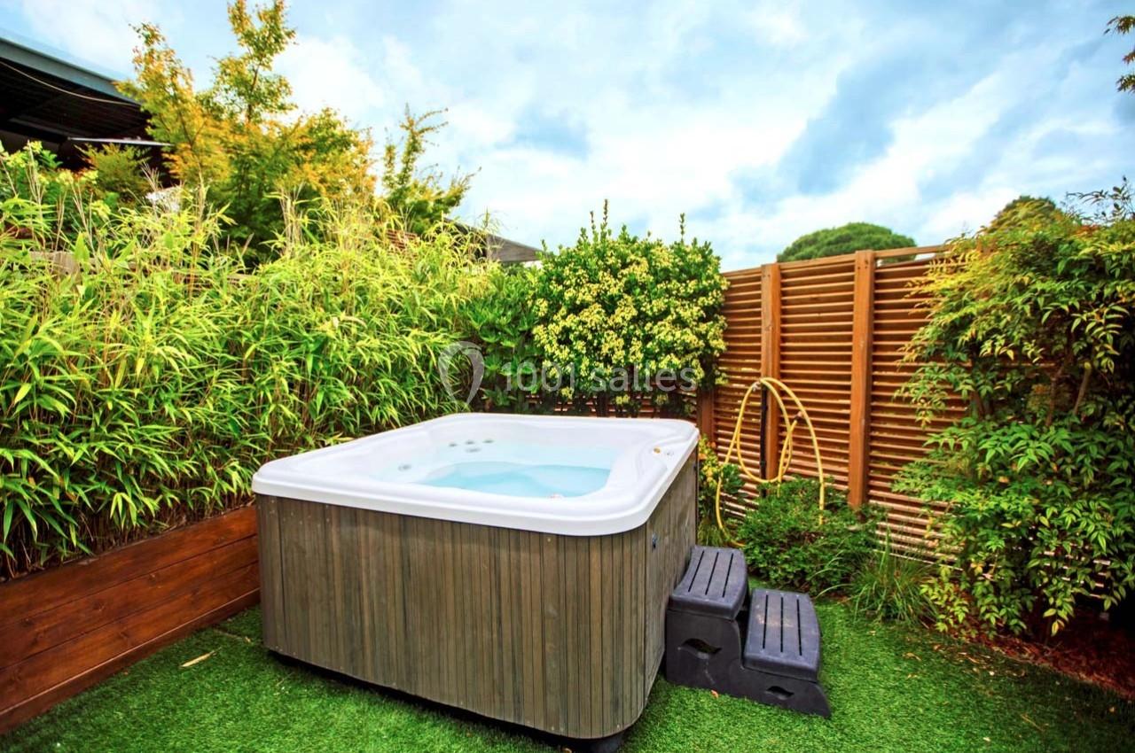 Spa extérieur entouré de plantes et de bambous, installé sur une pelouse synthétique avec des marches d'accès.