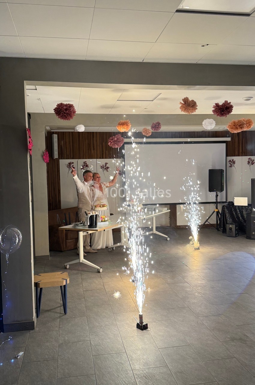 Un couple célèbre un événement dans une salle décorée, avec des étincelles lumineuses au sol et des guirlandes suspendues.