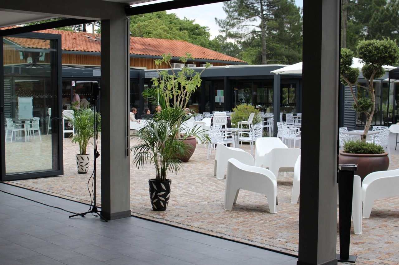 Vue depuis l'intérieur d'un bâtiment sur une terrasse avec des chaises blanches, des plantes en pot et un sol pavé.