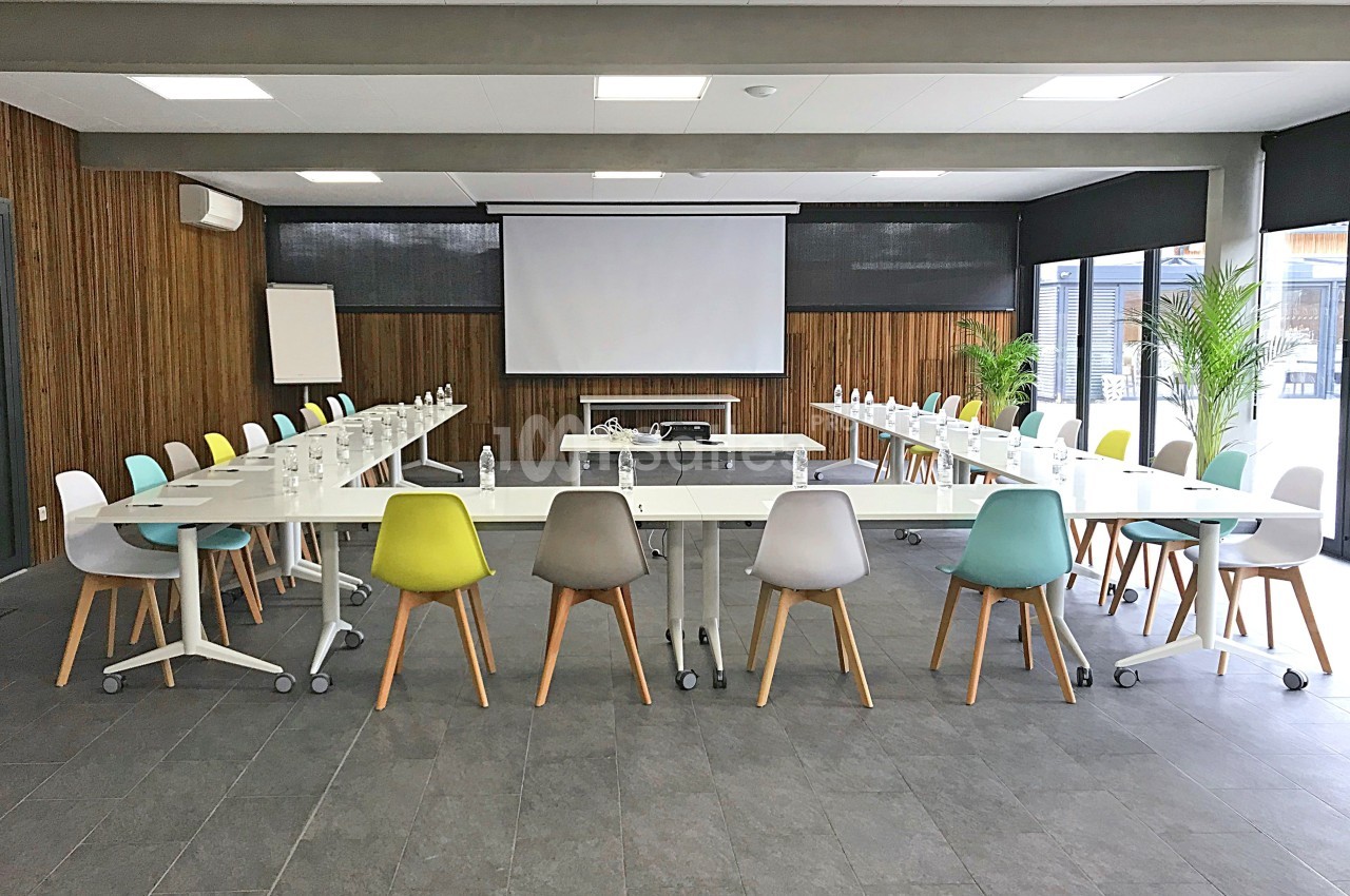 Salle de réunion lumineuse avec tables en U, chaises colorées, écran de projection et plantes en arrière-plan.