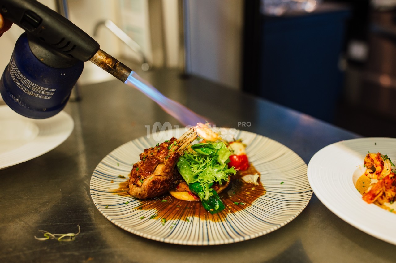 Un chef caramélise un plat de viande et légumes avec un chalumeau dans une cuisine professionnelle.
