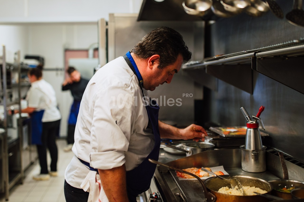 Un chef en tablier bleu prépare un plat dans une cuisine professionnelle, avec d'autres cuisiniers en arrière-plan.
