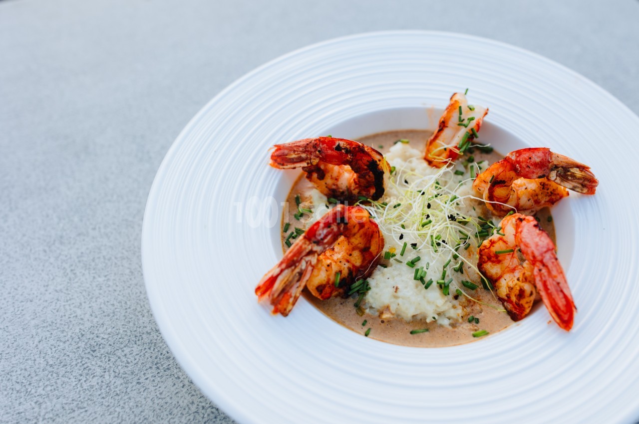 Assiette de crevettes grillées servies sur une purée avec une sauce et des pousses fraîches en garniture.