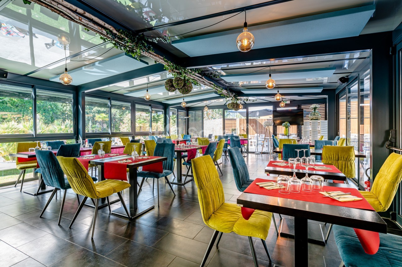 Salle de restaurant lumineuse avec tables dressées, chaises colorées et grandes baies vitrées donnant sur un espace…