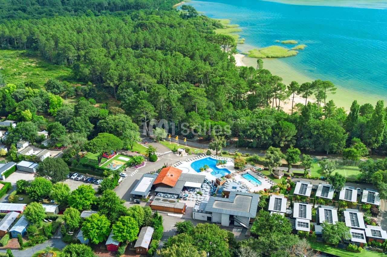 Vue aérienne d'un camping entouré de forêt, avec piscine, mobil-homes et accès à un lac bordé de végétation.