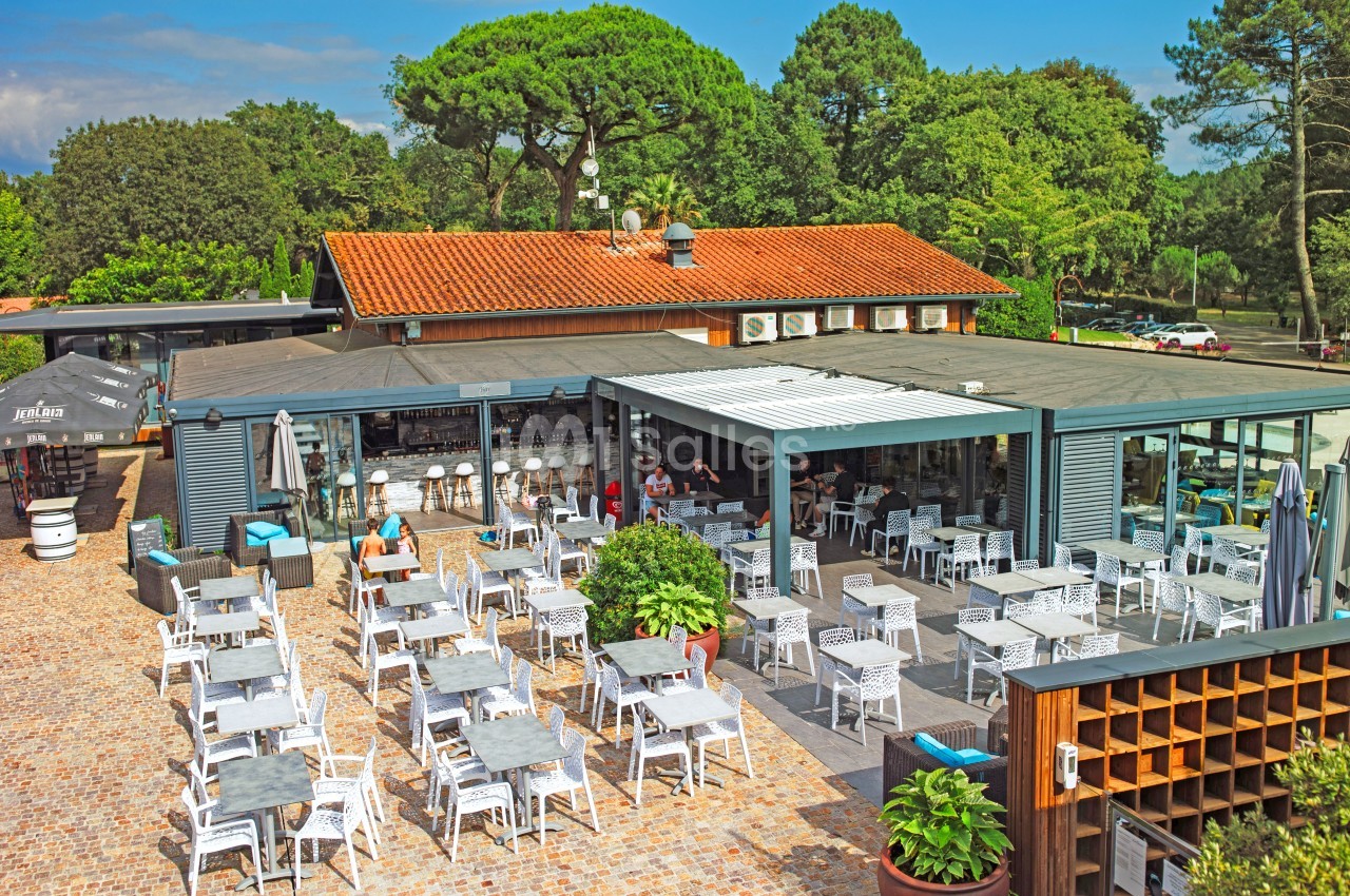 Terrasse d'un restaurant avec tables et chaises en extérieur, entourée de verdure et d'un bâtiment à toit rouge.