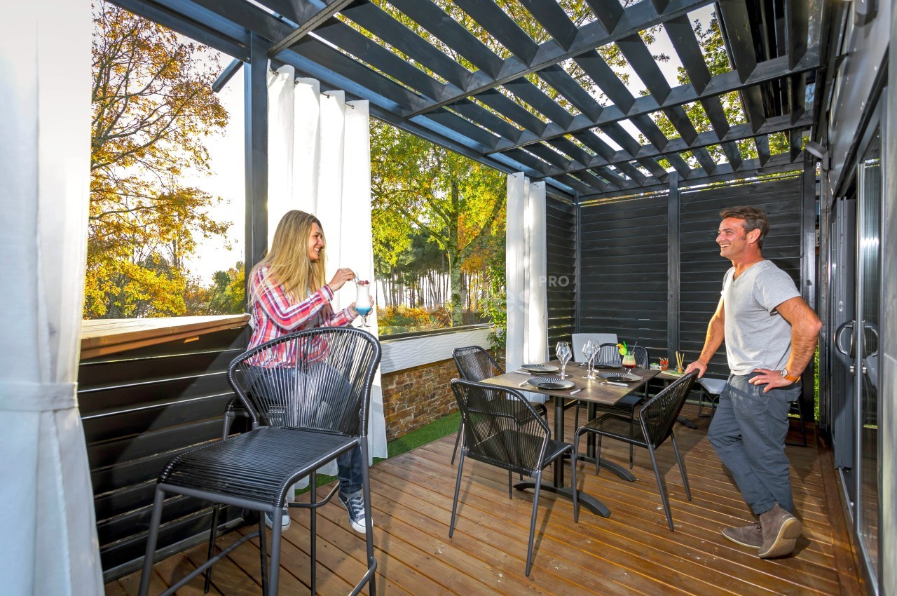Une femme et un homme discutent sur une terrasse en bois aménagée avec une table dressée et des chaises hautes.