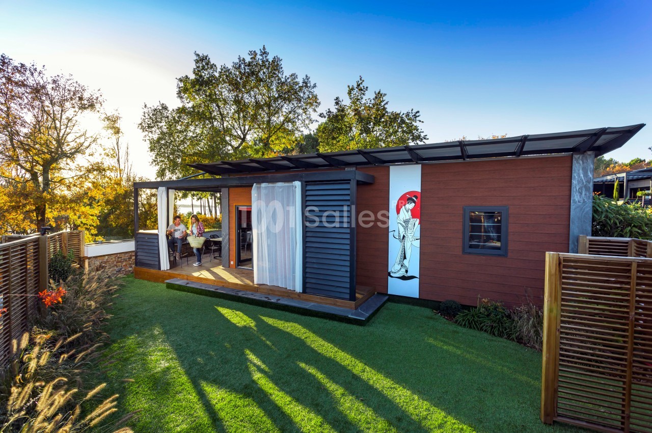 Maison modulaire en bois avec terrasse, située dans un jardin verdoyant sous un ciel ensoleillé.