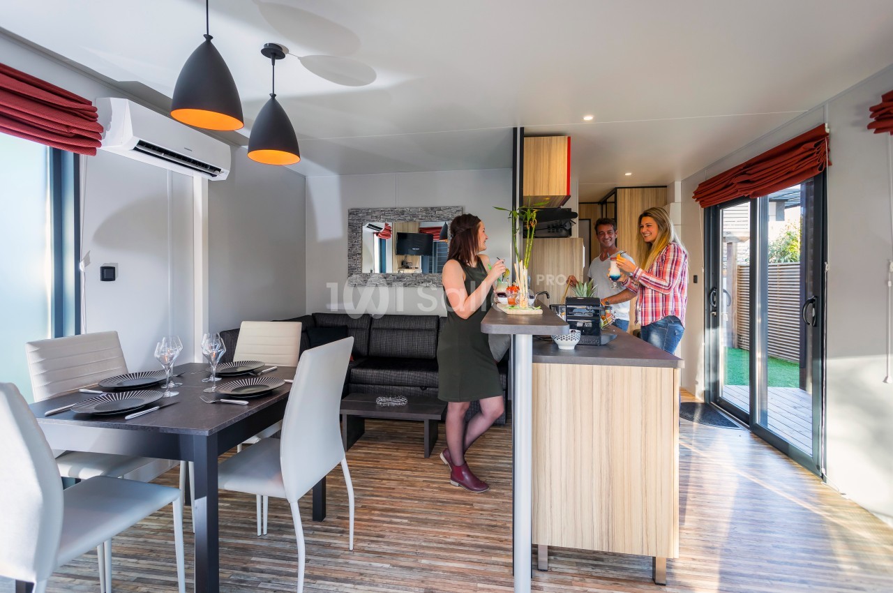 Trois personnes discutent dans une cuisine ouverte moderne avec table à manger et grandes baies vitrées.