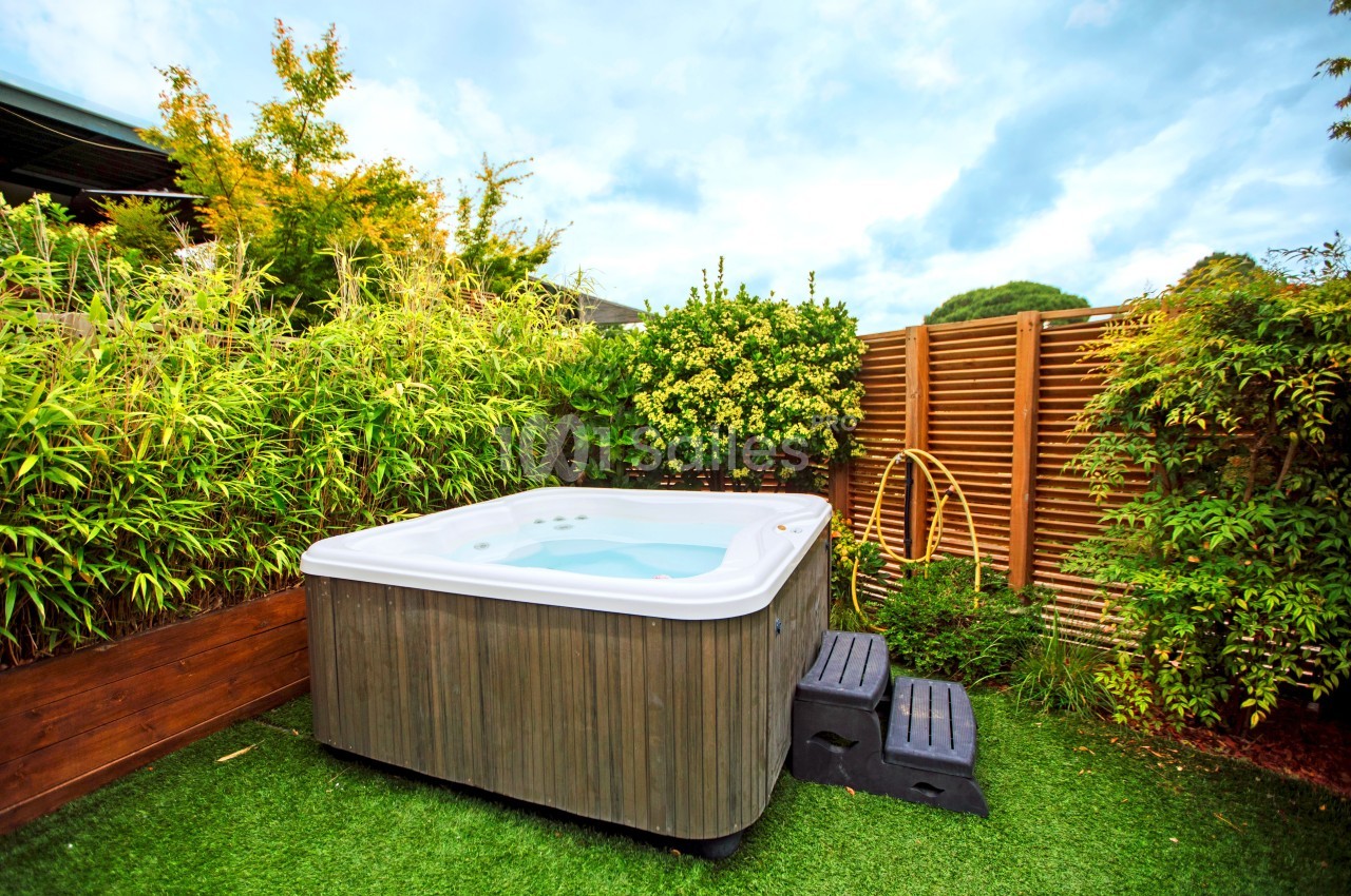 Spa extérieur entouré de plantes et de panneaux en bois, installé sur une pelouse synthétique avec des marches noires.