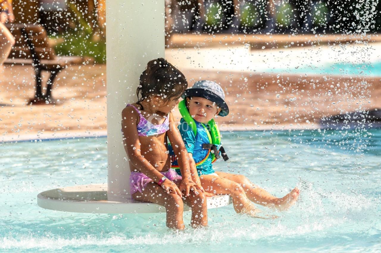 Deux enfants assis sous une fontaine d'eau dans une piscine peu profonde, portant des maillots de bain et des accessoires de…