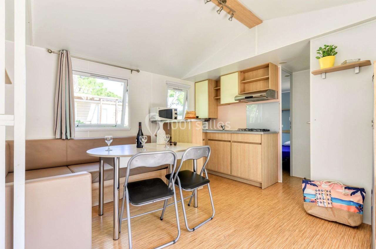 Intérieur d'un mobil-home avec coin cuisine équipé, table, chaises, banquette et lumière naturelle.