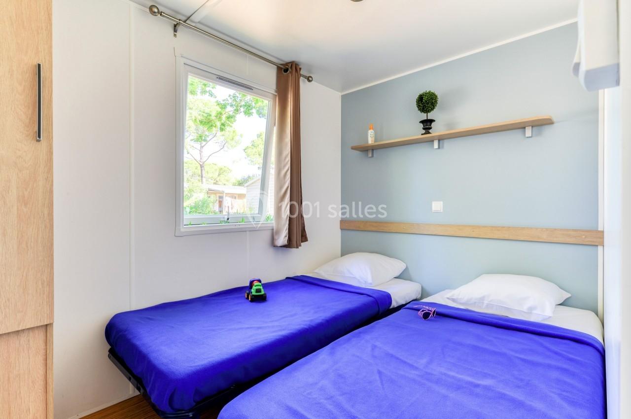 Deux lits simples avec draps bleus dans une chambre lumineuse, une étagère murale et une fenêtre donnant sur des arbres.