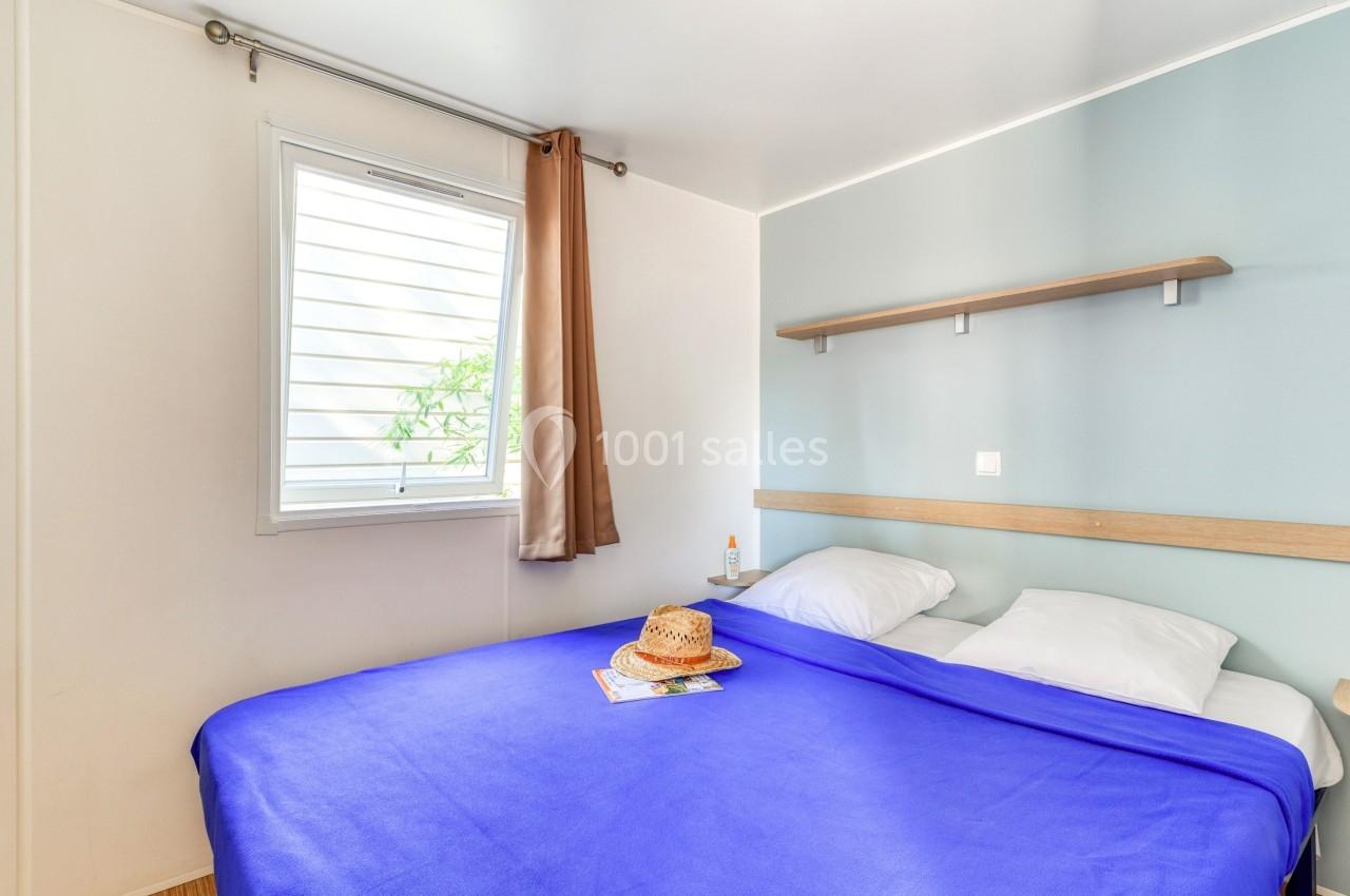 Chambre lumineuse avec un lit double, drap bleu, fenêtre avec rideau beige et étagère murale au-dessus du lit.