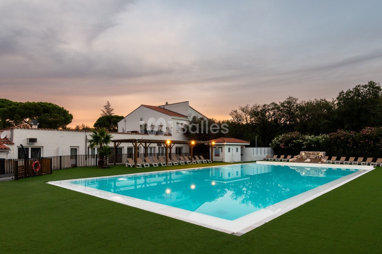 Piscine extérieure éclairée entourée de chaises longues, pelouse artificielle et bâtiments blancs au crépuscule.