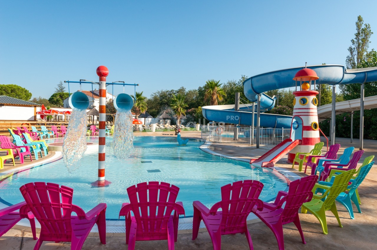 Piscine extérieure avec toboggans, jets d'eau et chaises colorées disposées autour sous un ciel dégagé.