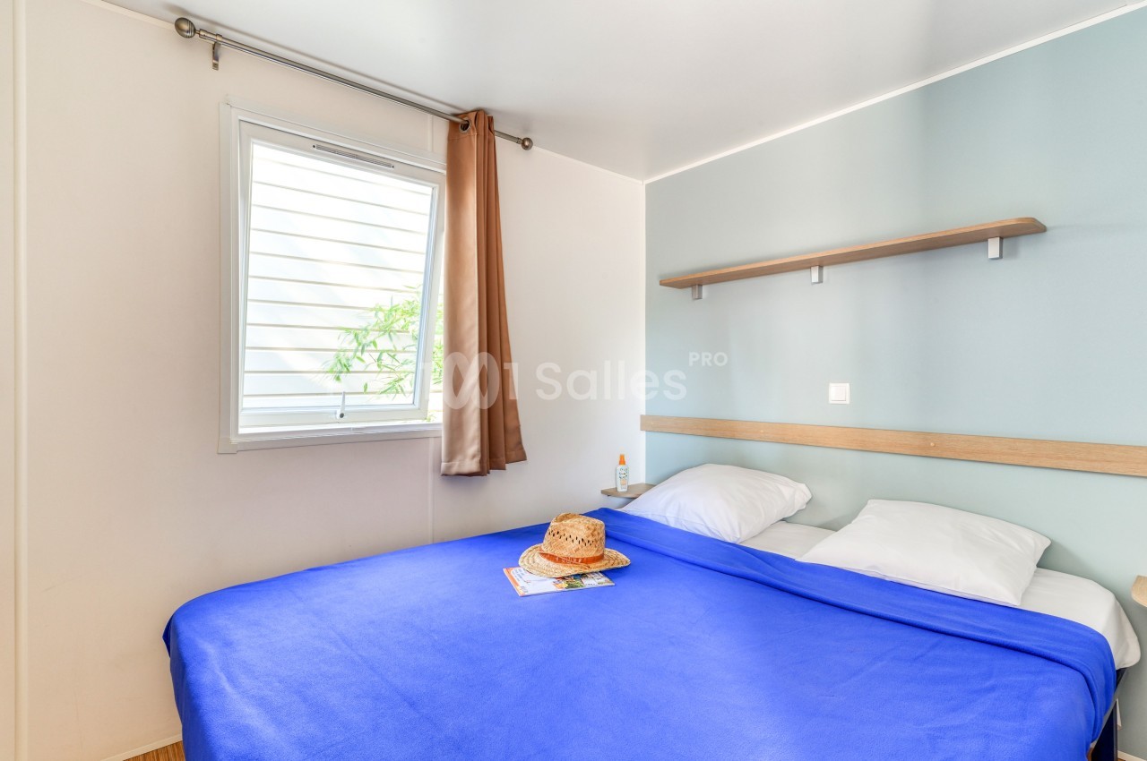 Chambre lumineuse avec un lit double, drap bleu, fenêtre avec rideau beige, étagère murale et chapeau posé sur le lit.
