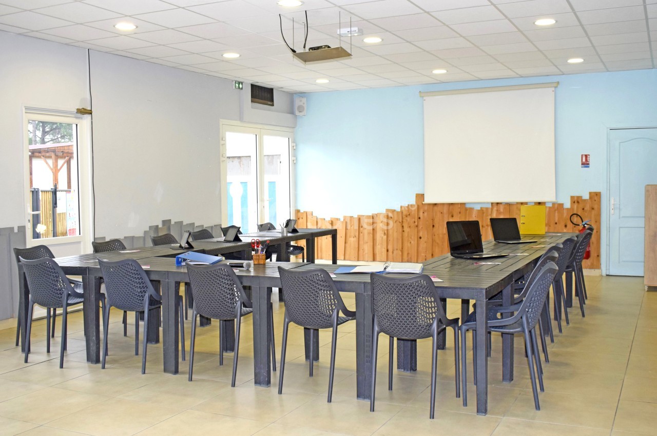 Salle de réunion lumineuse avec tables en U, chaises en plastique, projecteur au plafond et écran blanc.