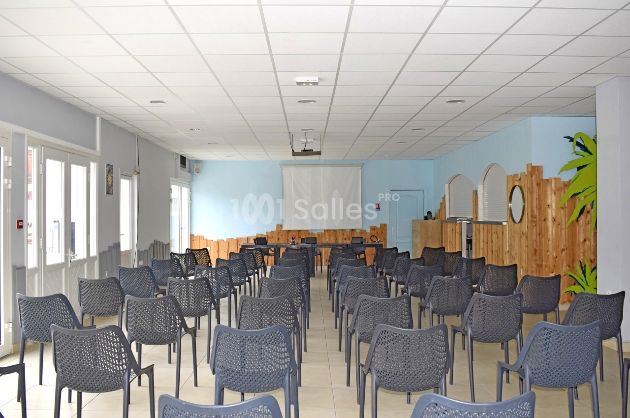 Salle lumineuse avec des rangées de chaises en plastique noir, un écran de projection et des murs décorés de bois clair.
