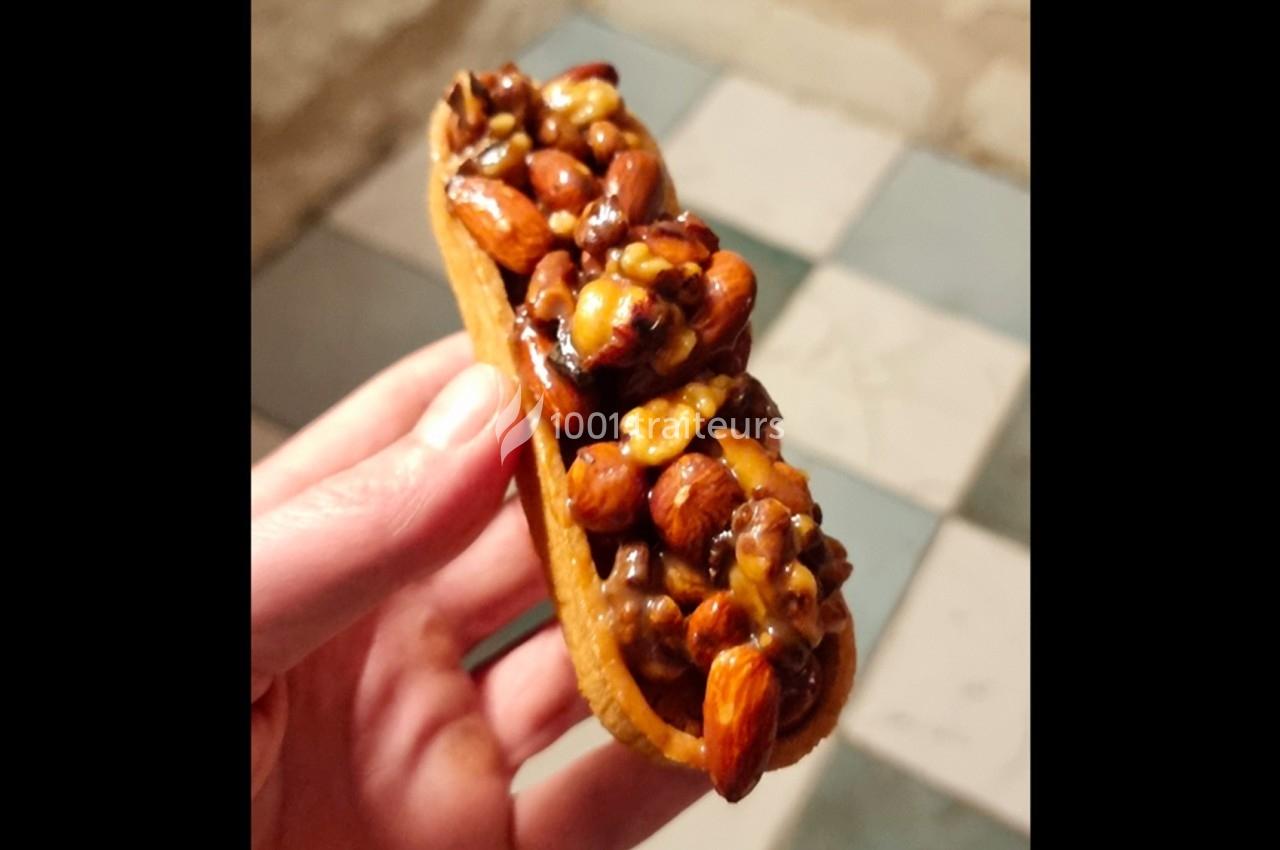 Tartelette garnie de noix, amandes et noisettes caramélisées, tenue dans une main sur un sol carrelé.