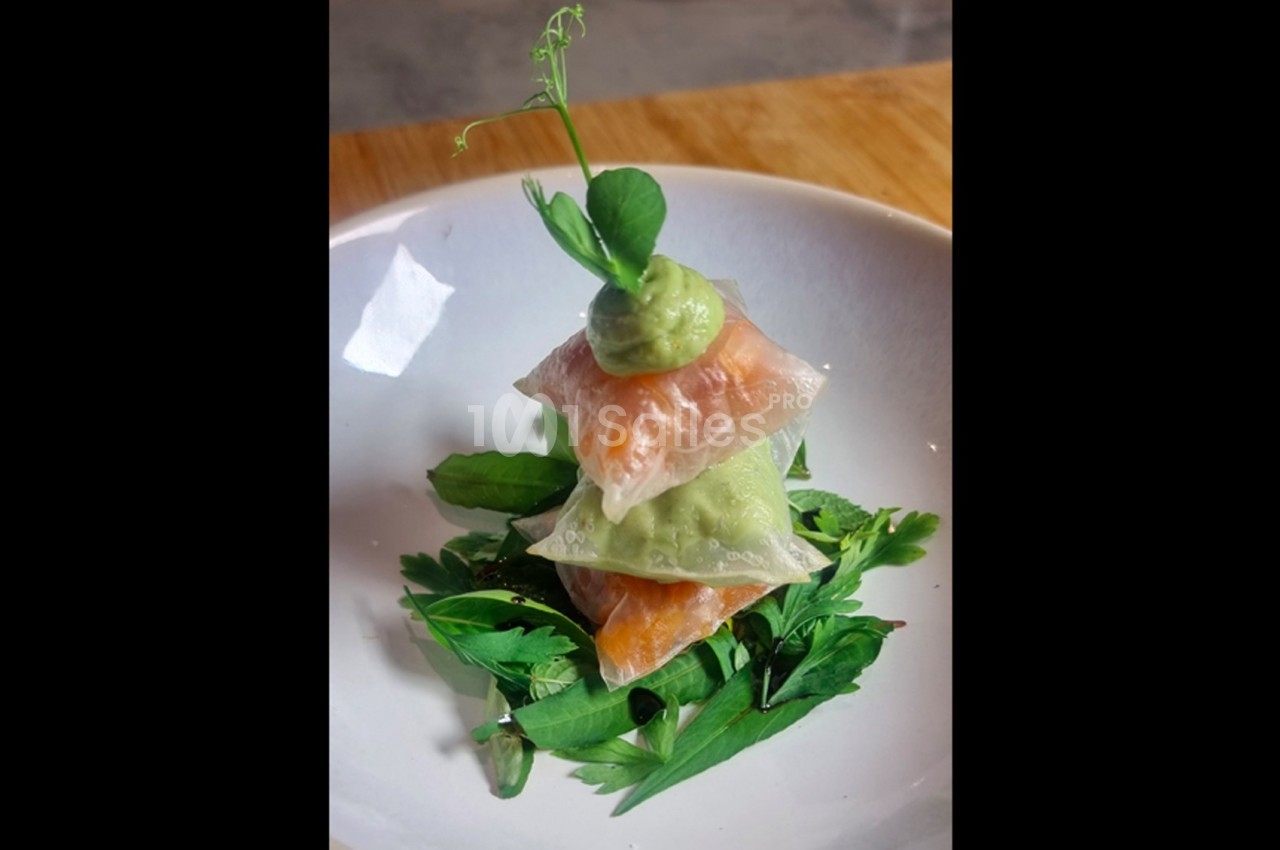 Empilement de ravioles transparentes garnies de saumon et d'avocat, décoré d'une pousse verte, sur un lit de verdure.