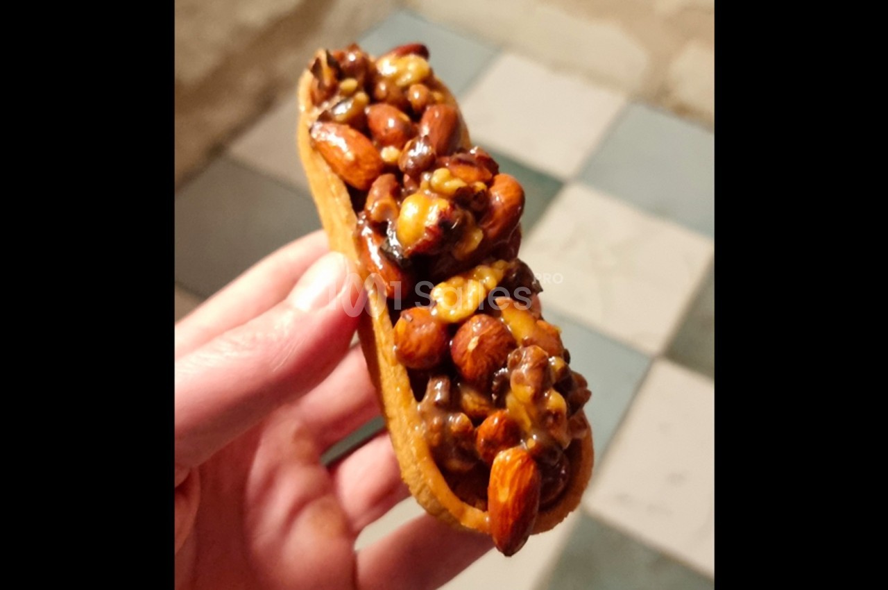 Tartelette garnie de noix, amandes et noisettes caramélisées, tenue dans une main sur un sol carrelé.
