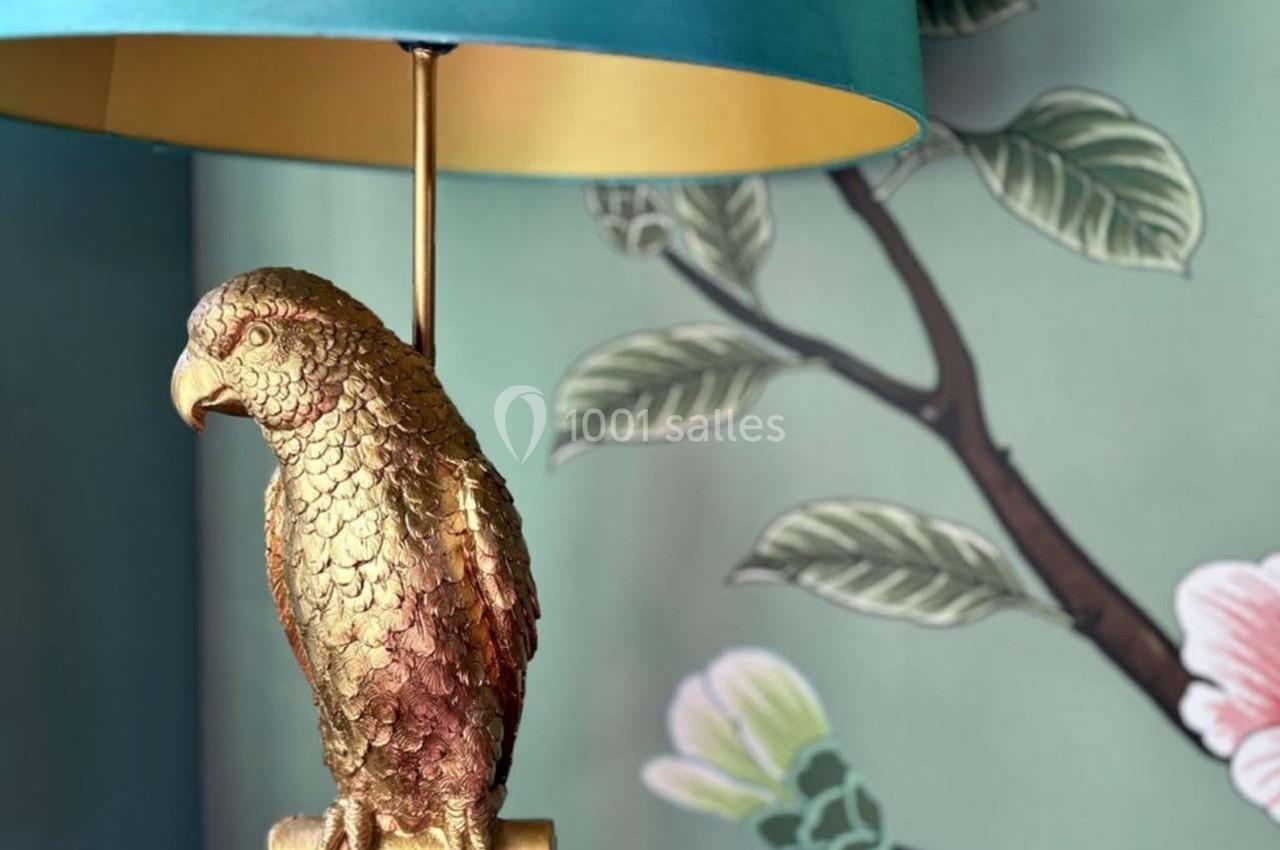 Lampe avec pied en forme de perroquet doré, abat-jour turquoise et fond mural à motifs floraux.