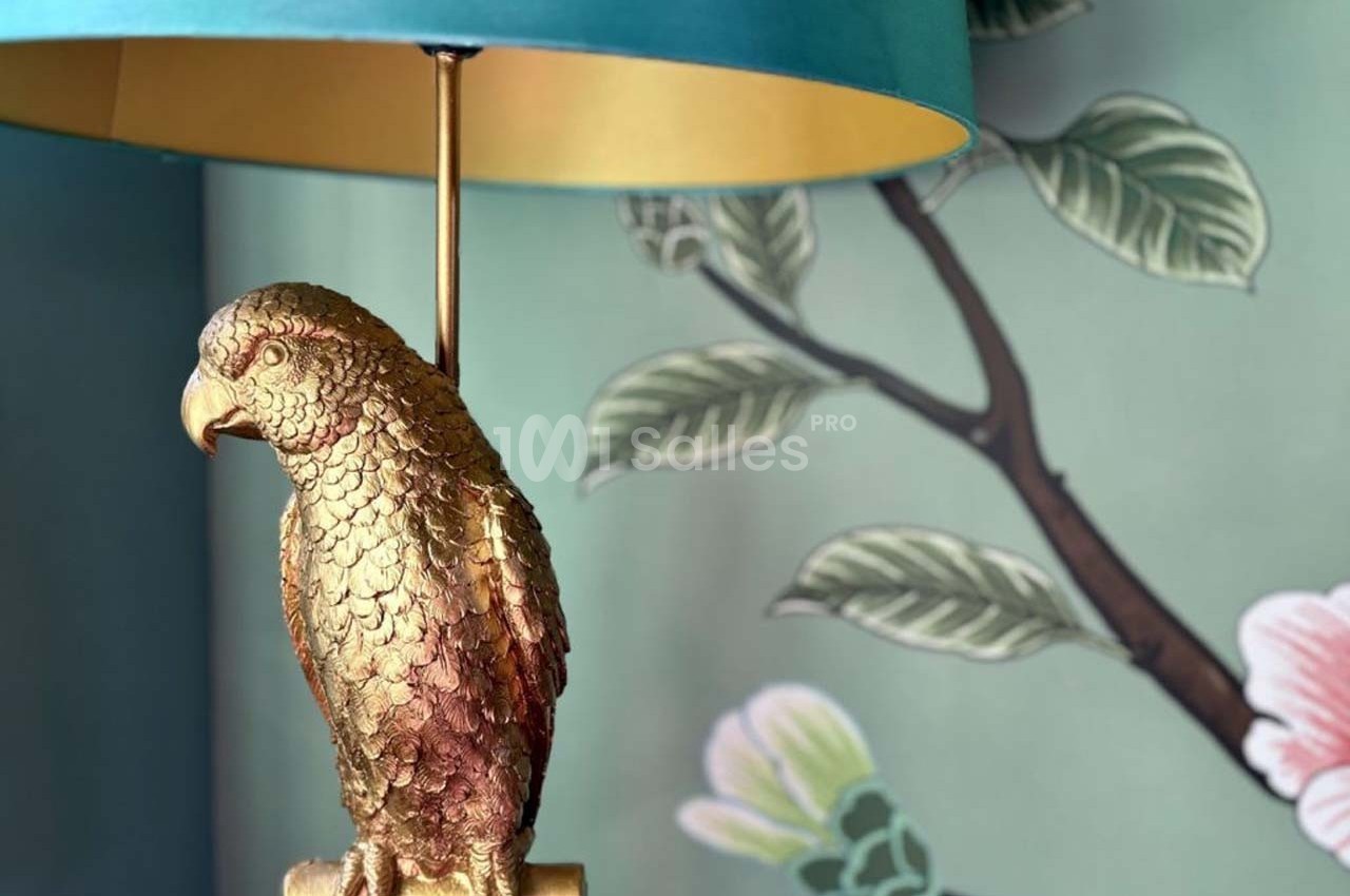 Lampe avec pied en forme de perroquet doré, posée devant un mur décoré de motifs floraux et feuillages.