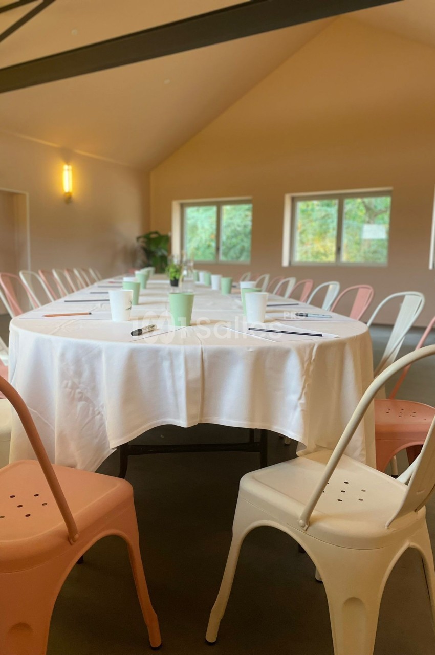 Salle lumineuse avec une table ronde dressée de nappes blanches, entourée de chaises pastel et équipée de carnets et…