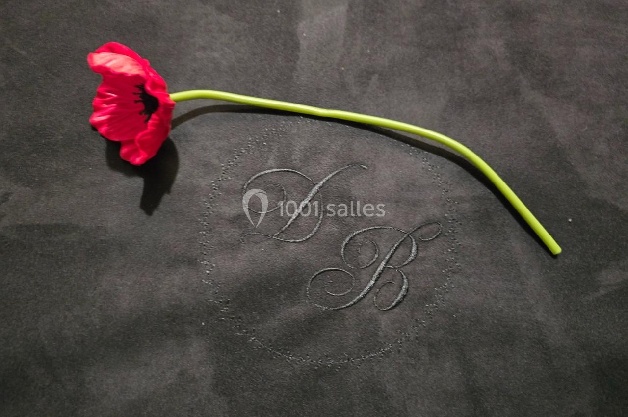 Fleur rouge à longue tige posée sur un tissu noir avec des initiales brodées en arrière-plan.
