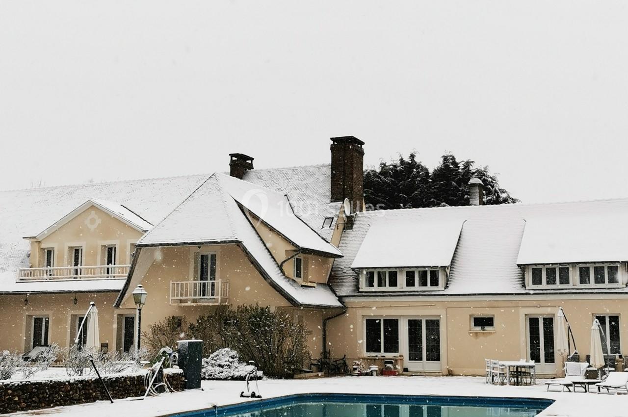 Grande maison beige avec piscine extérieure, entourée de neige sous un ciel gris.