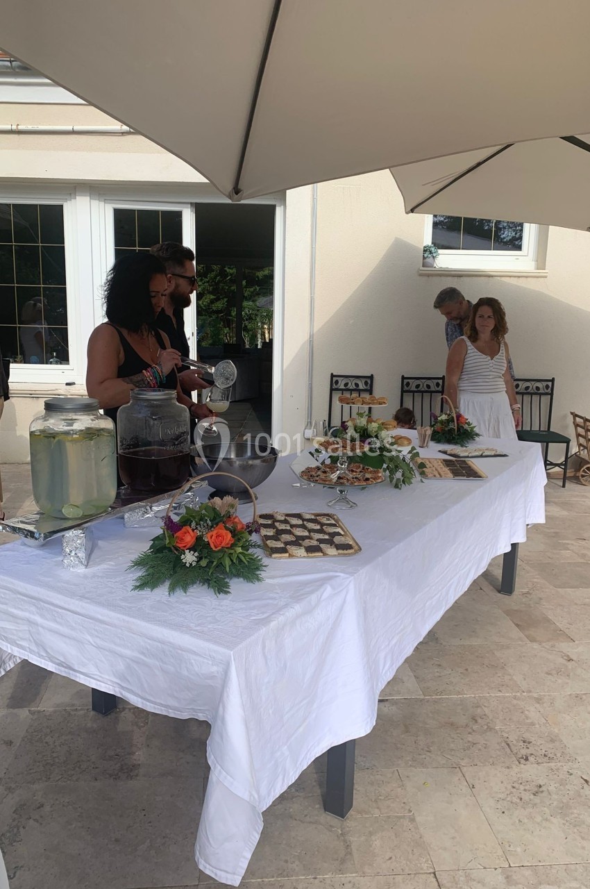 Buffet en extérieur avec boissons, desserts et décorations florales, des invités se servant sous un parasol.