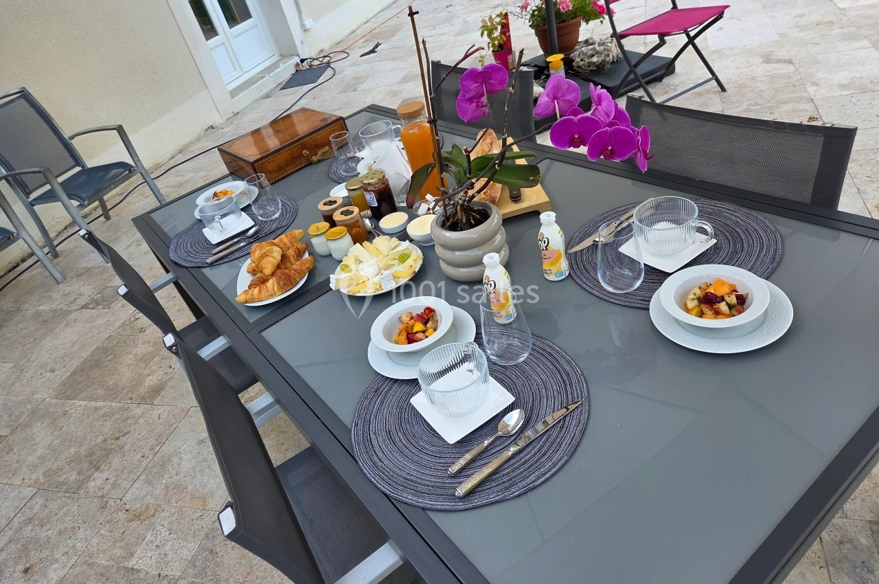Table de petit-déjeuner en extérieur avec croissants, fruits, fromage, jus d'orange et orchidées en décoration.