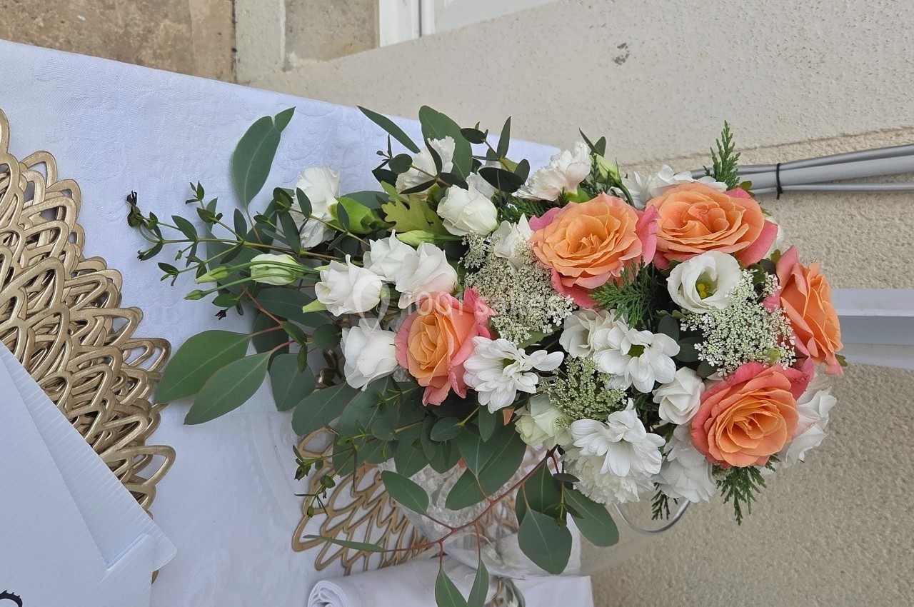 Bouquet de fleurs composé de roses orange, fleurs blanches et feuillage vert, posé sur une nappe blanche.