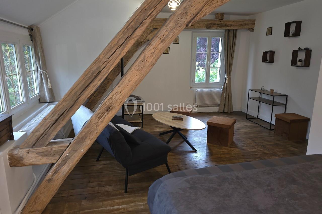 Pièce mansardée avec poutres apparentes, canapé, table basse, lit et vue sur un jardin par une fenêtre.