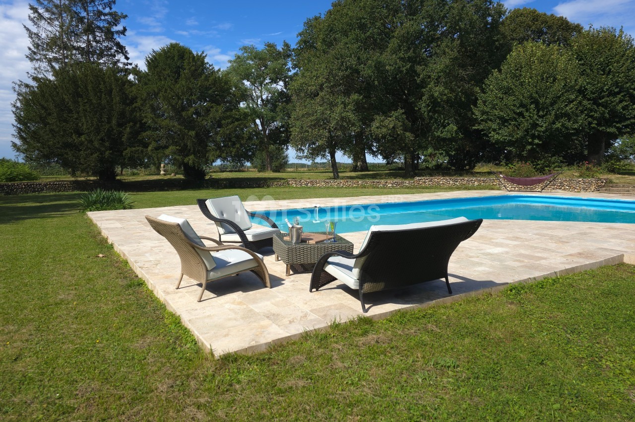 Mobilier de jardin disposé près d'une piscine entourée de pelouse et d'arbres sous un ciel dégagé.