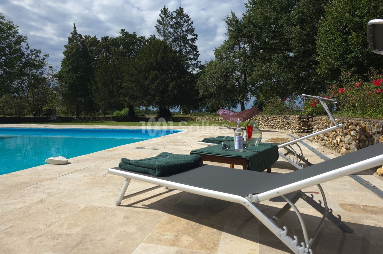 Deux transats près d'une piscine, avec une table dressée contenant des verres et une bouteille, entourée de végétation.