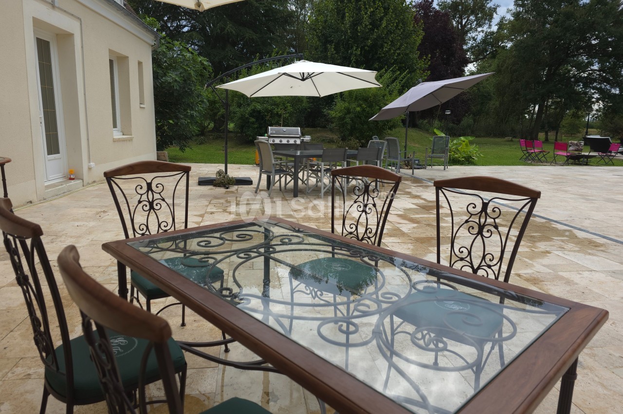Table en verre avec chaises en métal sur une terrasse en pierre, entourée de parasols et d'un jardin verdoyant.