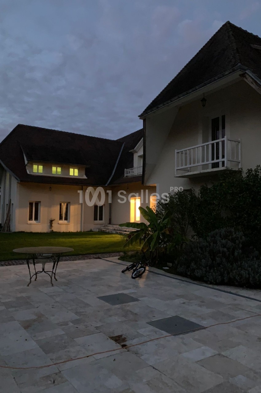 Maison avec toit en pente, éclairée de l'intérieur, vue depuis une terrasse en pierre au crépuscule.