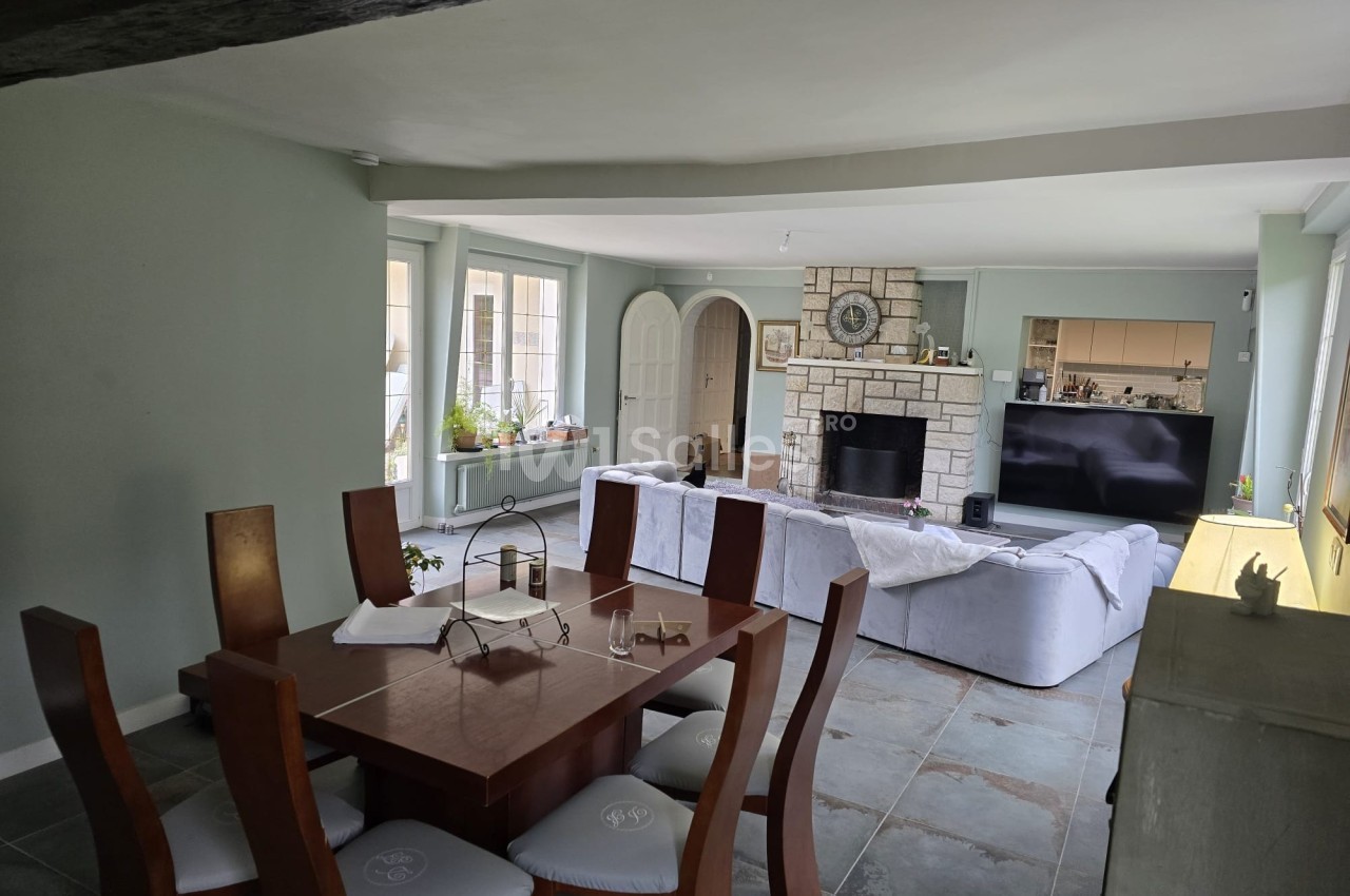 Salle à manger avec table en bois, chaises rembourrées, salon avec canapé, cheminée en pierre et télévision.