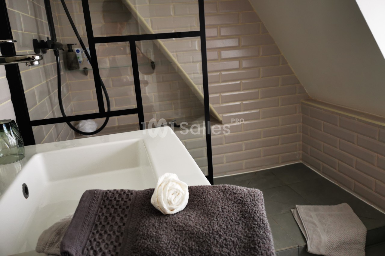 Salle de bain mansardée avec lavabo, serviettes pliées, douche à paroi vitrée et carrelage beige.