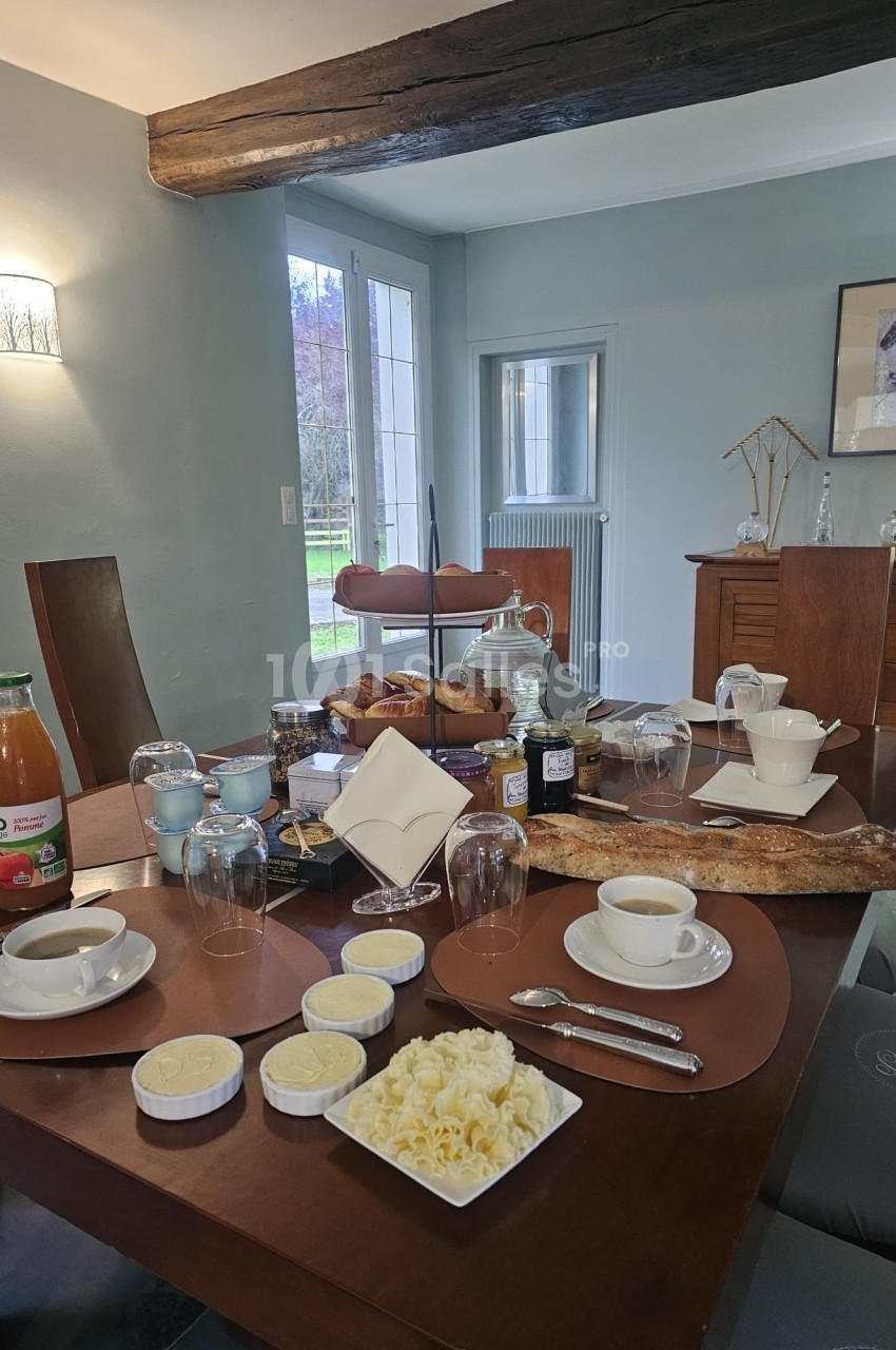 Table dressée pour un petit-déjeuner avec pain, viennoiseries, confitures, jus de fruit, café et vaisselle.